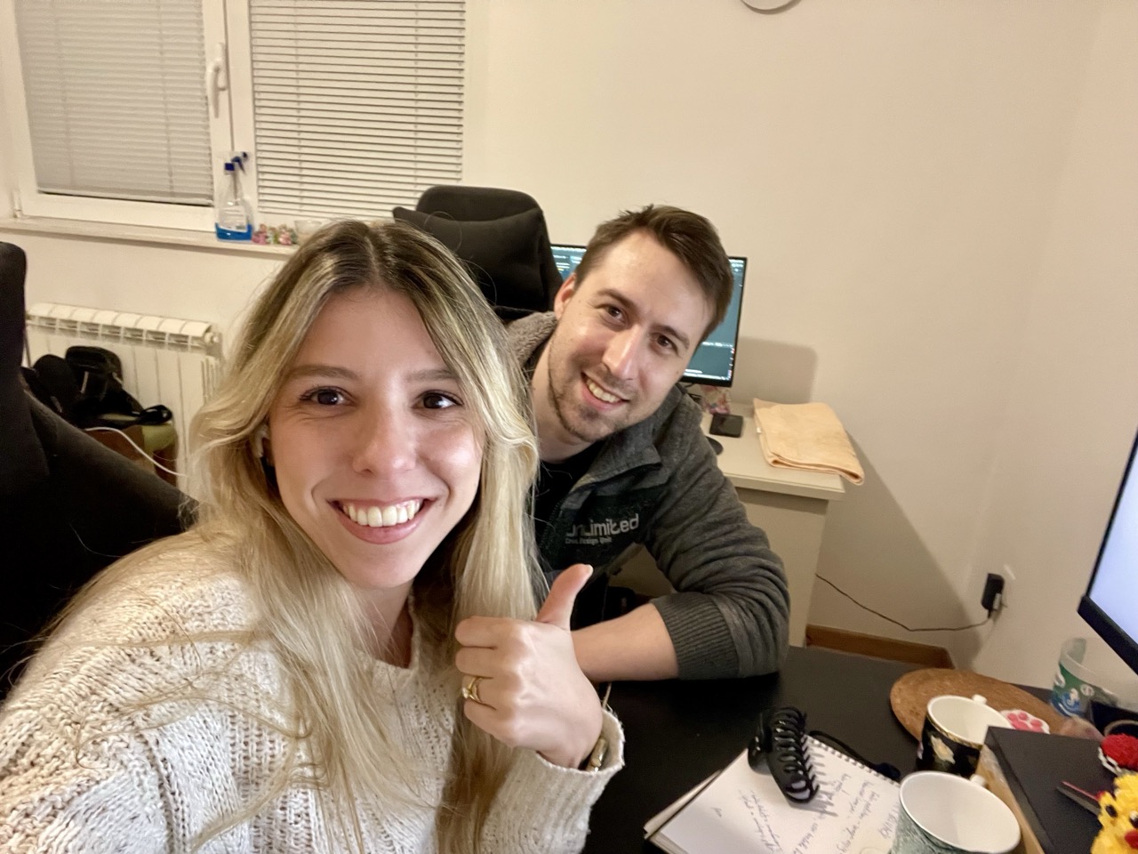 A man and woman in their living room. The woman is giving a thumbs up. Computers in the background.