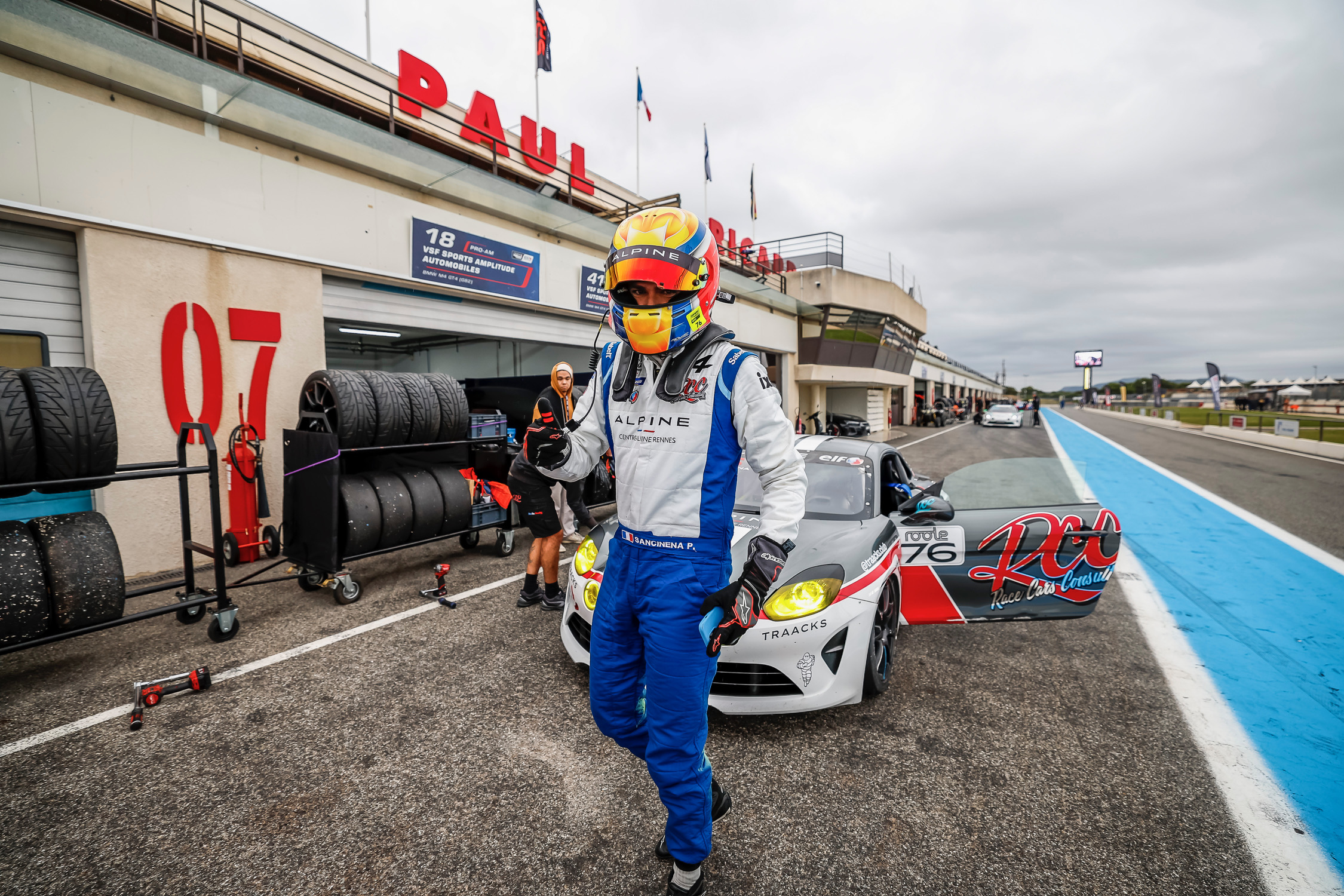 A man in a race suit gives a thumbs up to the camera. In the background is a race car and a race track. 