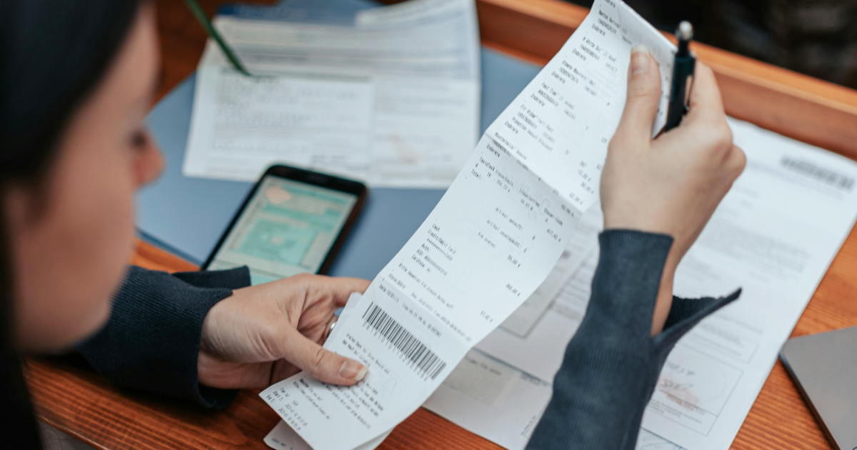 Person manually unrolling and reviewing a long paper receipt alongside other paperwork and a phone, the receipt keeping app problem