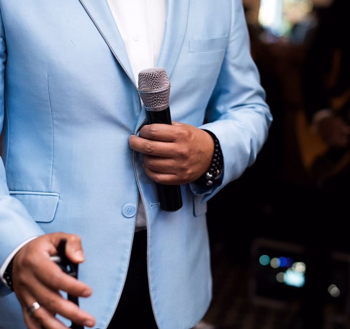 A close-up of a person in a light blue blazer holding a microphone, with a black wrist bracelet visible, set against a blurred background.