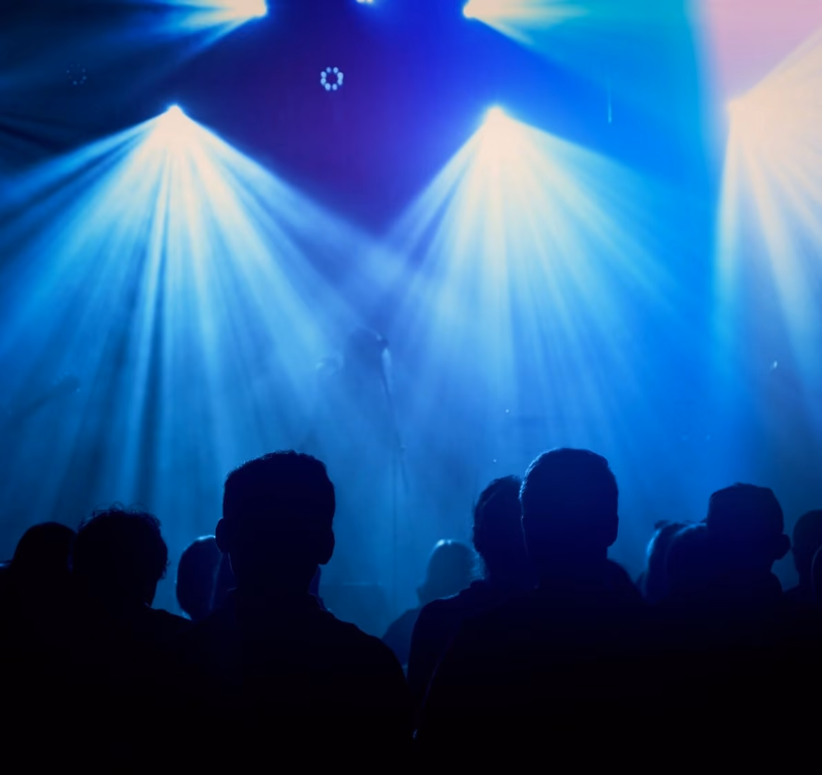 Silhouettes of an audience enjoying a live performance, illuminated by striking blue and white stage lights in a smoky atmosphere.