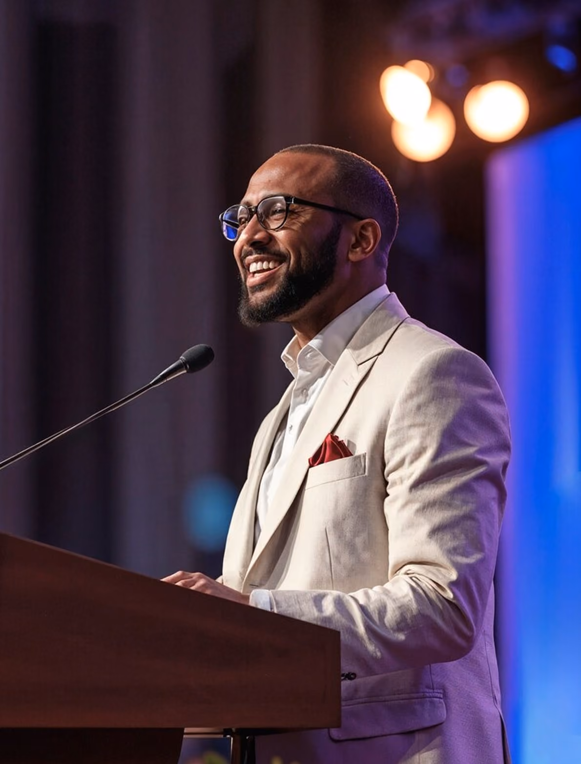 A well-dressed speaker stands at a podium, gesturing passionately during a presentation under warm stage lights.