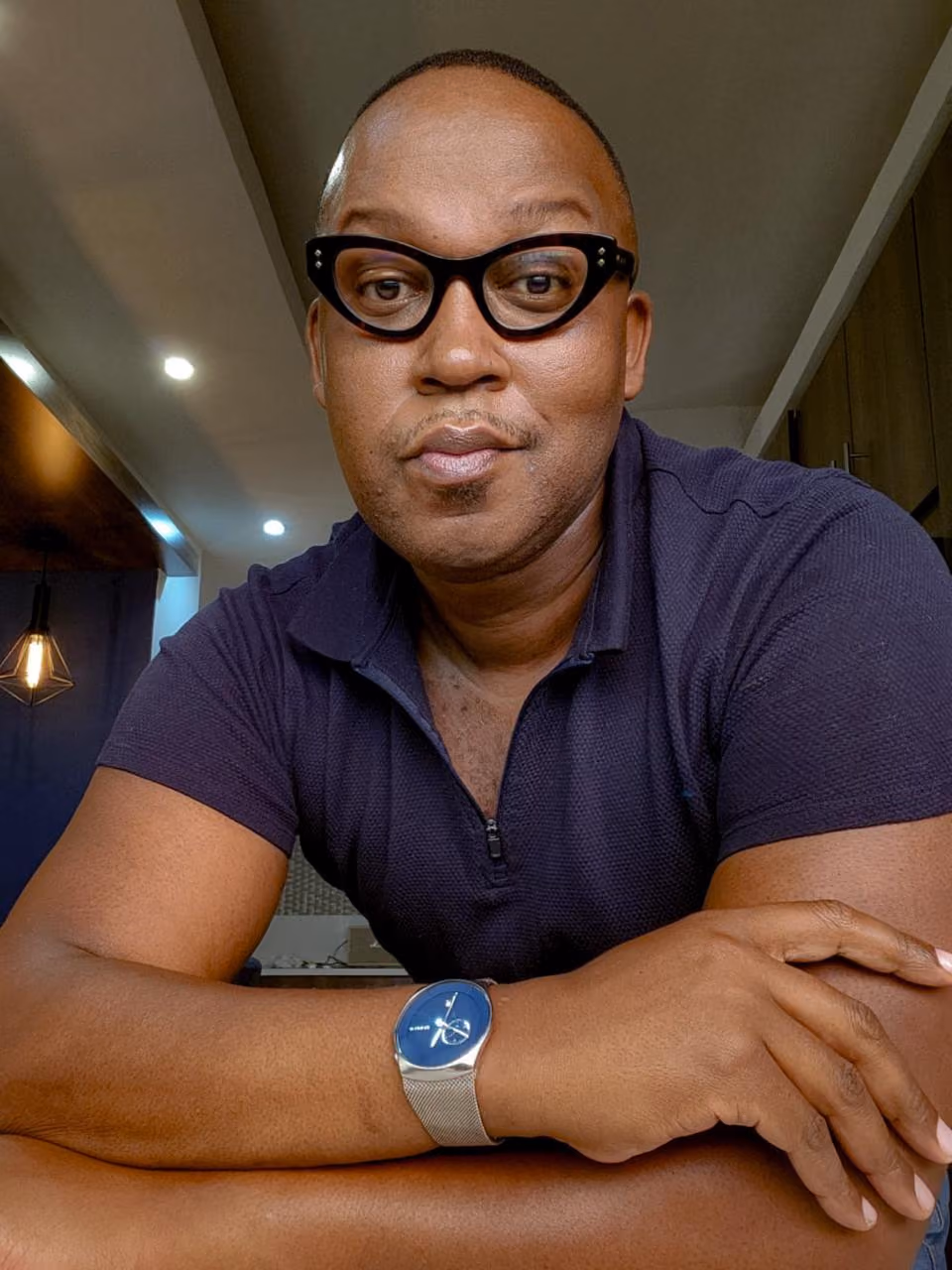 Man with short hair wearing black thick-rimmed glasses, a dark purple short-sleeve shirt, and a silver wristwatch with a blue face, sitting indoors with arms crossed on a table, looking at the camera.