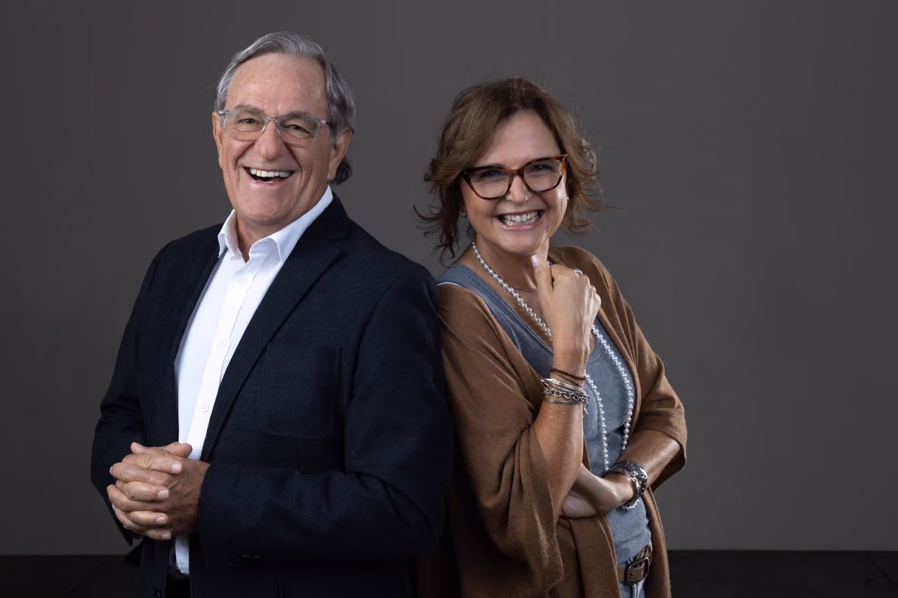 Smiling mature man in suit and woman in glasses stand back to back against gray background.