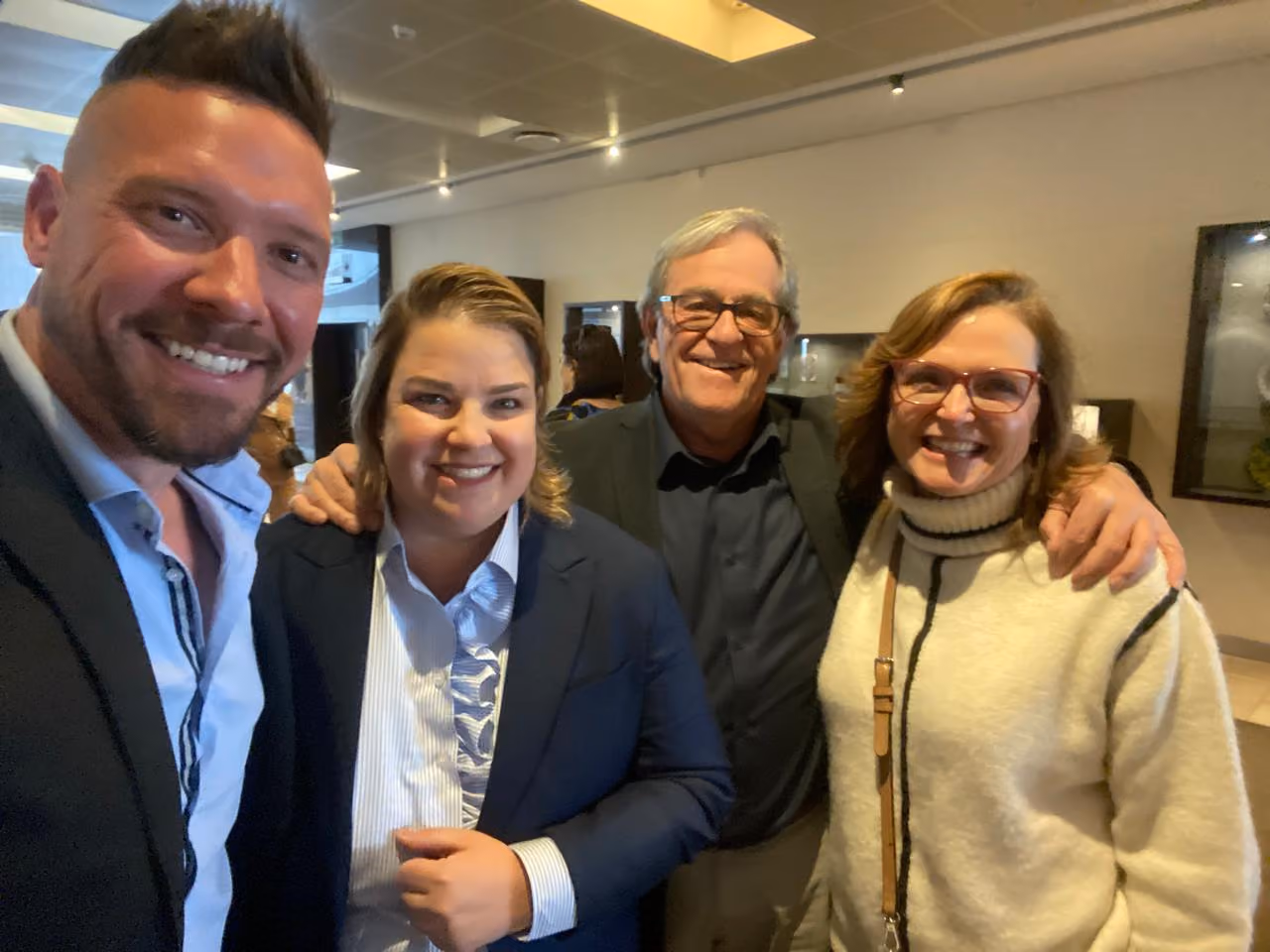 Four smiling people posing closely indoors, two men and two women, wearing glasses and formal to casual clothes.