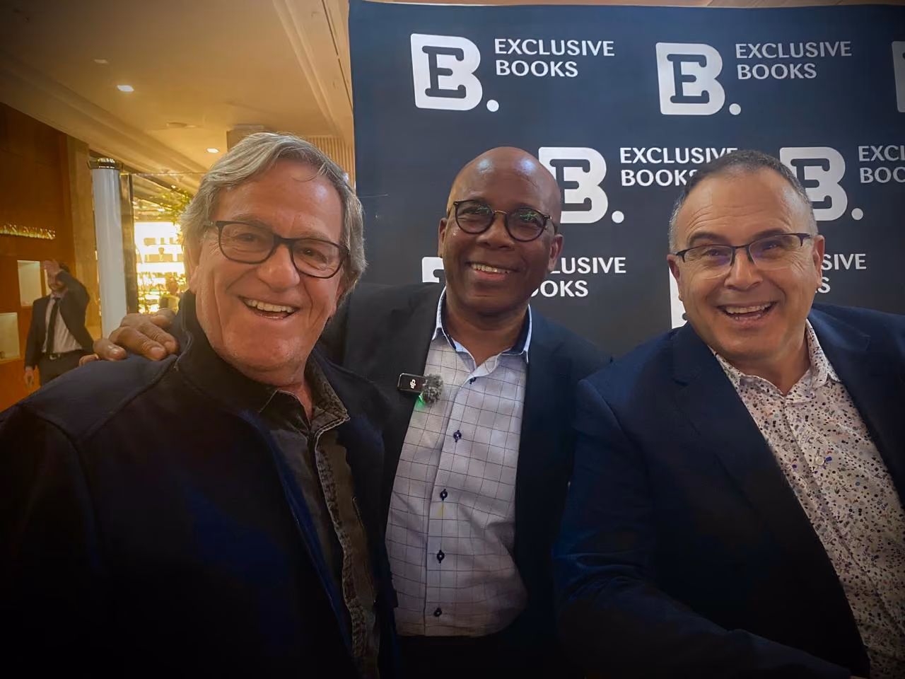 Three men smiling and posing in front of an Exclusive Books backdrop at a book event.