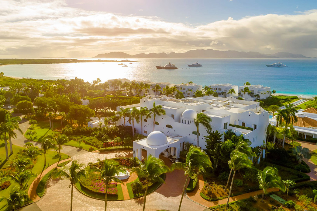 alt="Vue aérienne d’un hôtel de luxe aux toits blancs entouré de palmiers face à la mer turquoise à Anguilla dans les Caraïbes