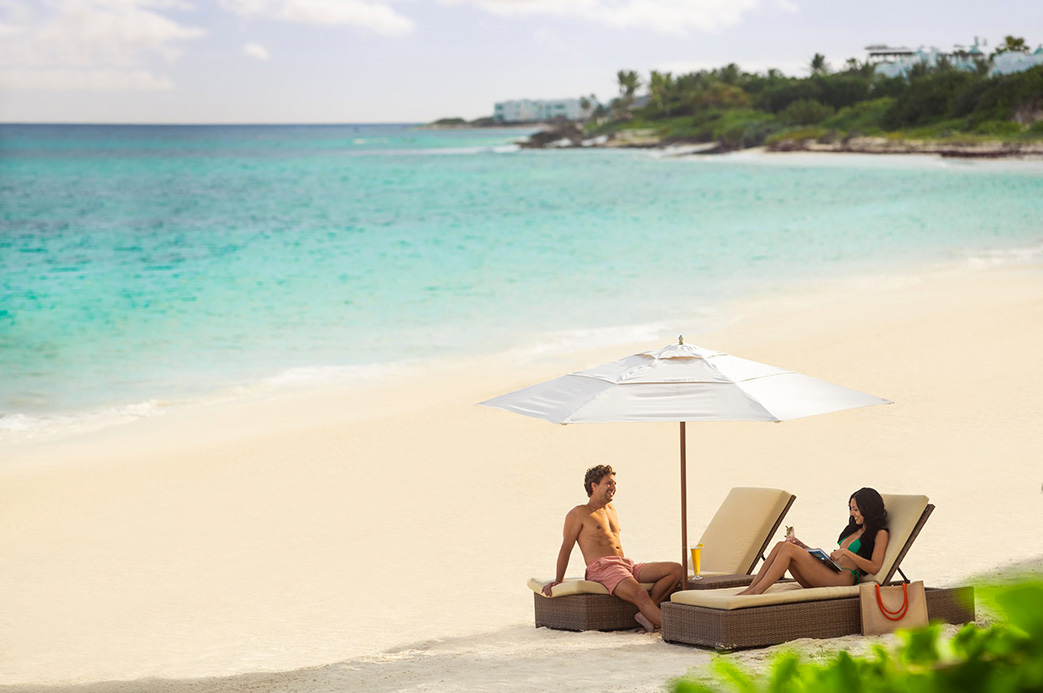 Couple se détendant sur des transats sous un parasol blanc sur une plage de sable fin aux Caraïbes, face à une mer turquoise
