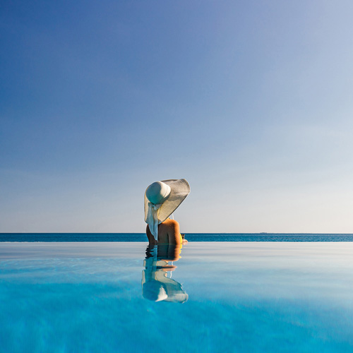 A woman wearing a hat is sitting in the middle of an infinity pool overlooking the sea.