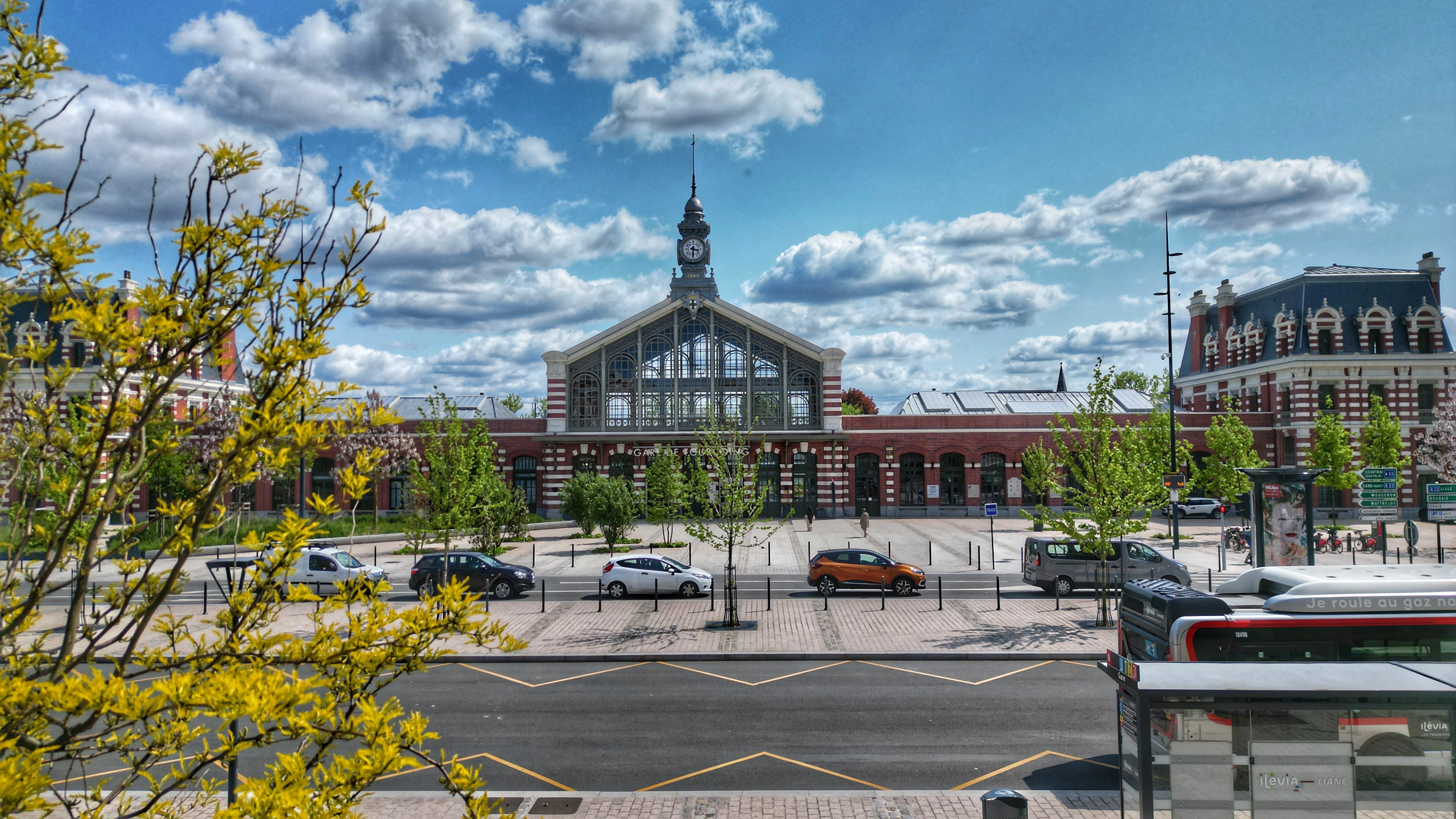 Vue extérieure de la gare de Tourcoing avec son bâtiment en briques rouges et blanches, une horloge centrale, des voitures et un bus arrêtés devant, arbres et arbustes au premier plan sous un ciel bleu avec quelques nuages.