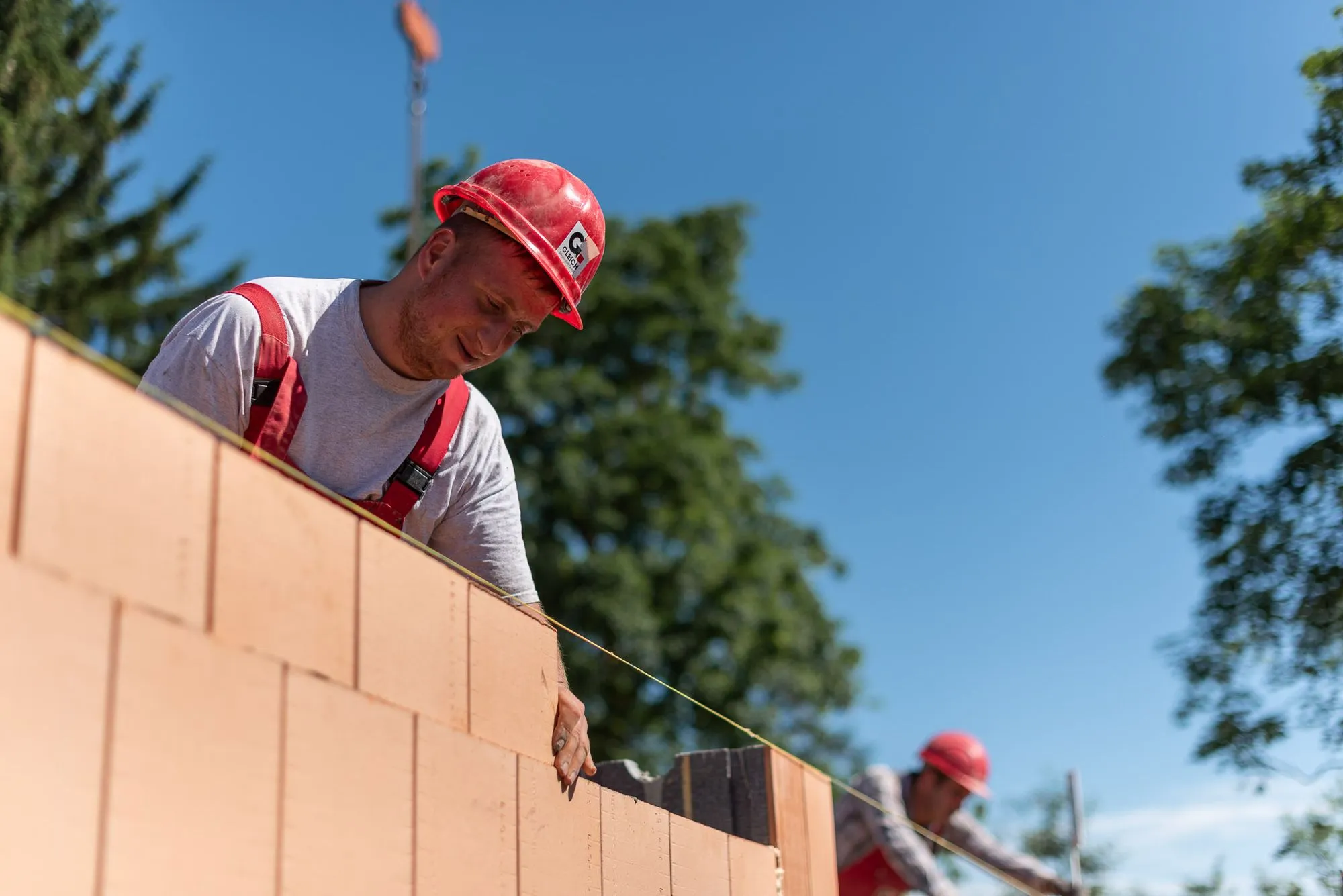 Bauarbeiter mit rotem Helm und roten Hosenträgern misst mit einer Schnur eine Mauer aus Backsteinen bei sonnigem Himmel.