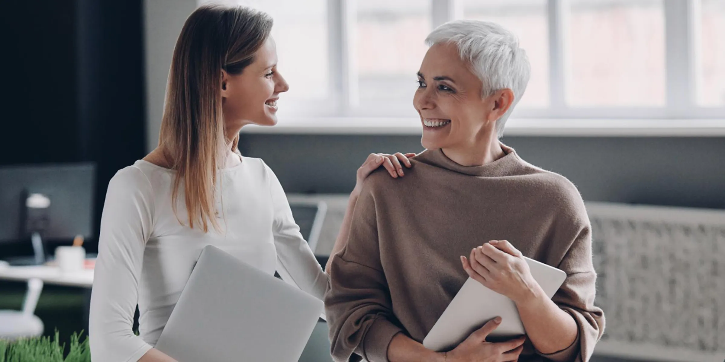 Two professional women smiling and connecting in a modern office setting.