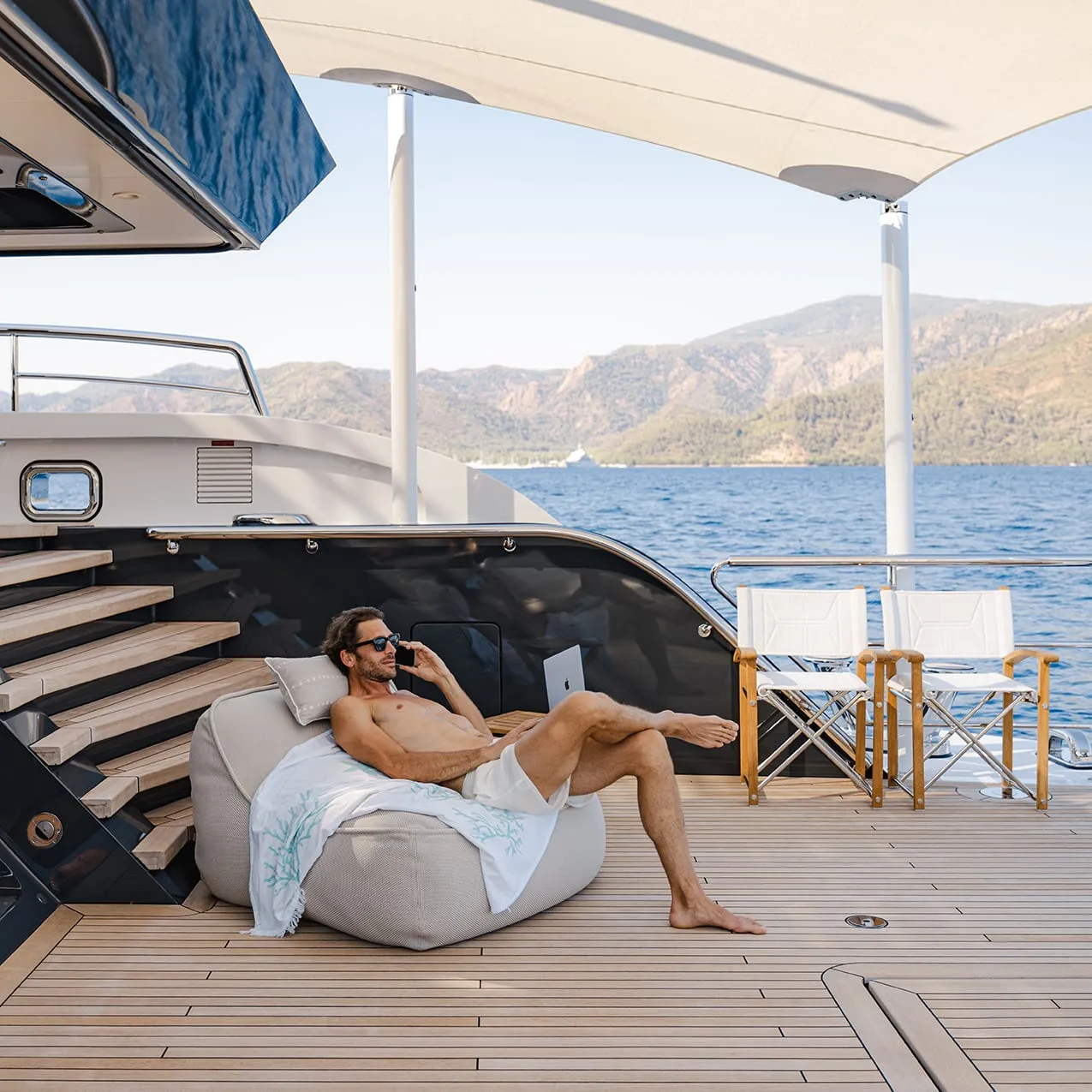 Man relaxing on a luxury yacht deck, talking on the phone with a laptop, with sea and mountain view in the background