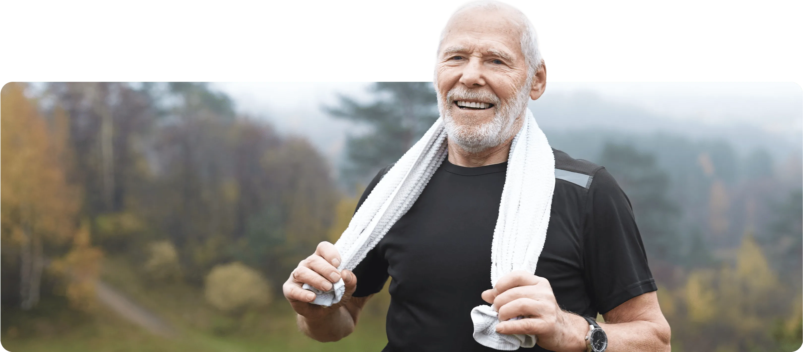 Smiling elderly man wearing black athletic shirt and white towel around neck outdoors in forest area.