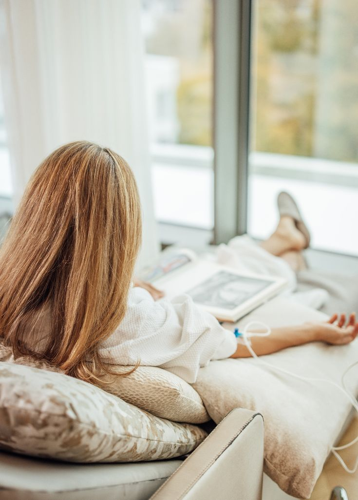 Person with light brown hair reclines on a couch reading a magazine near a window.