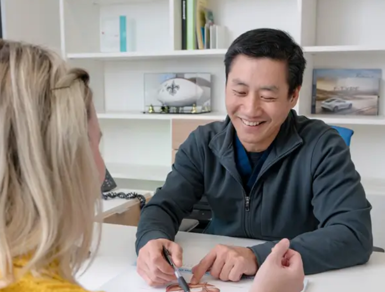 Smiling man in a dark jacket talking with a blonde woman across a desk in an office.