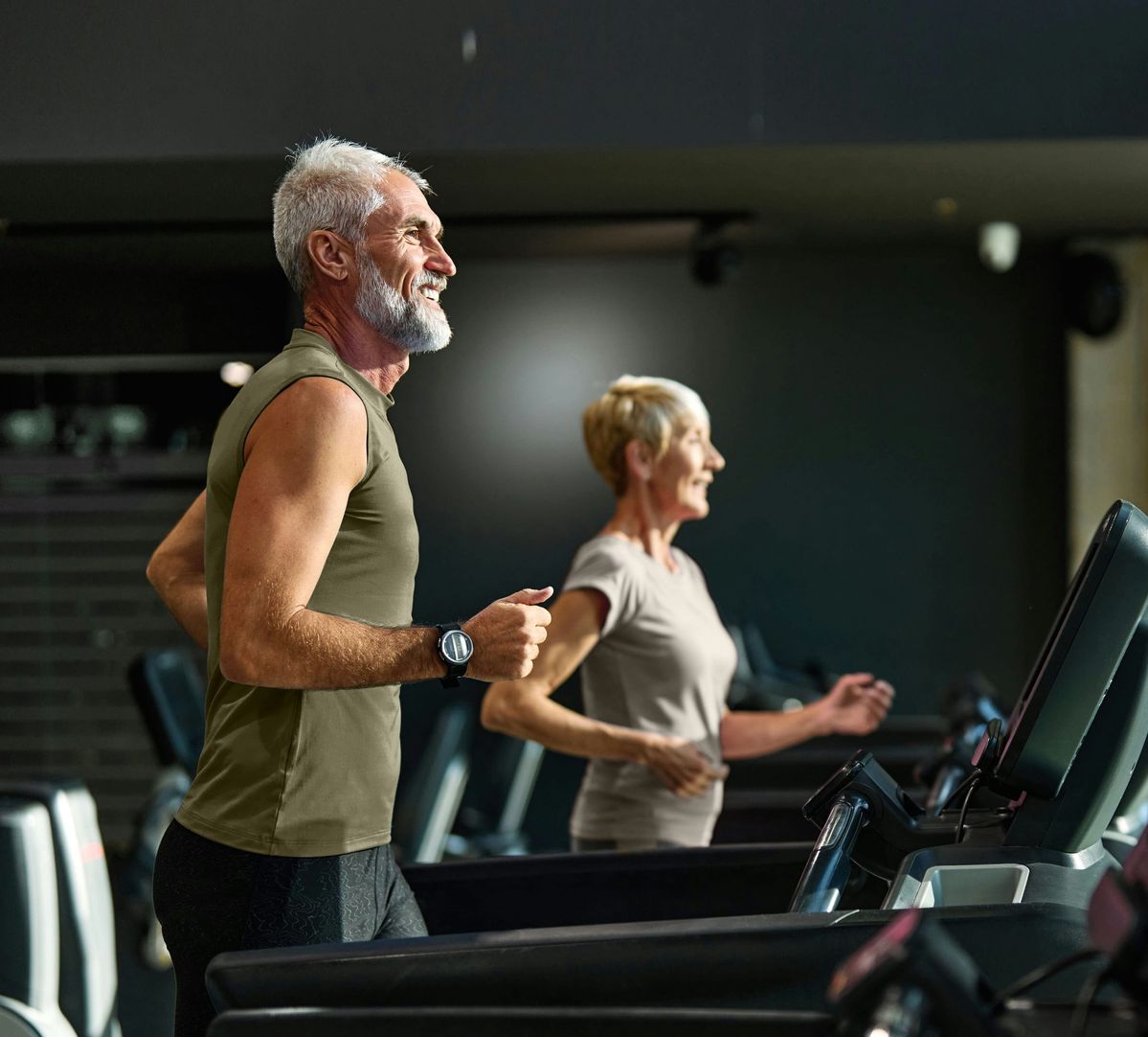 Smiling senior man and woman jogging side by side on treadmills in a gym.