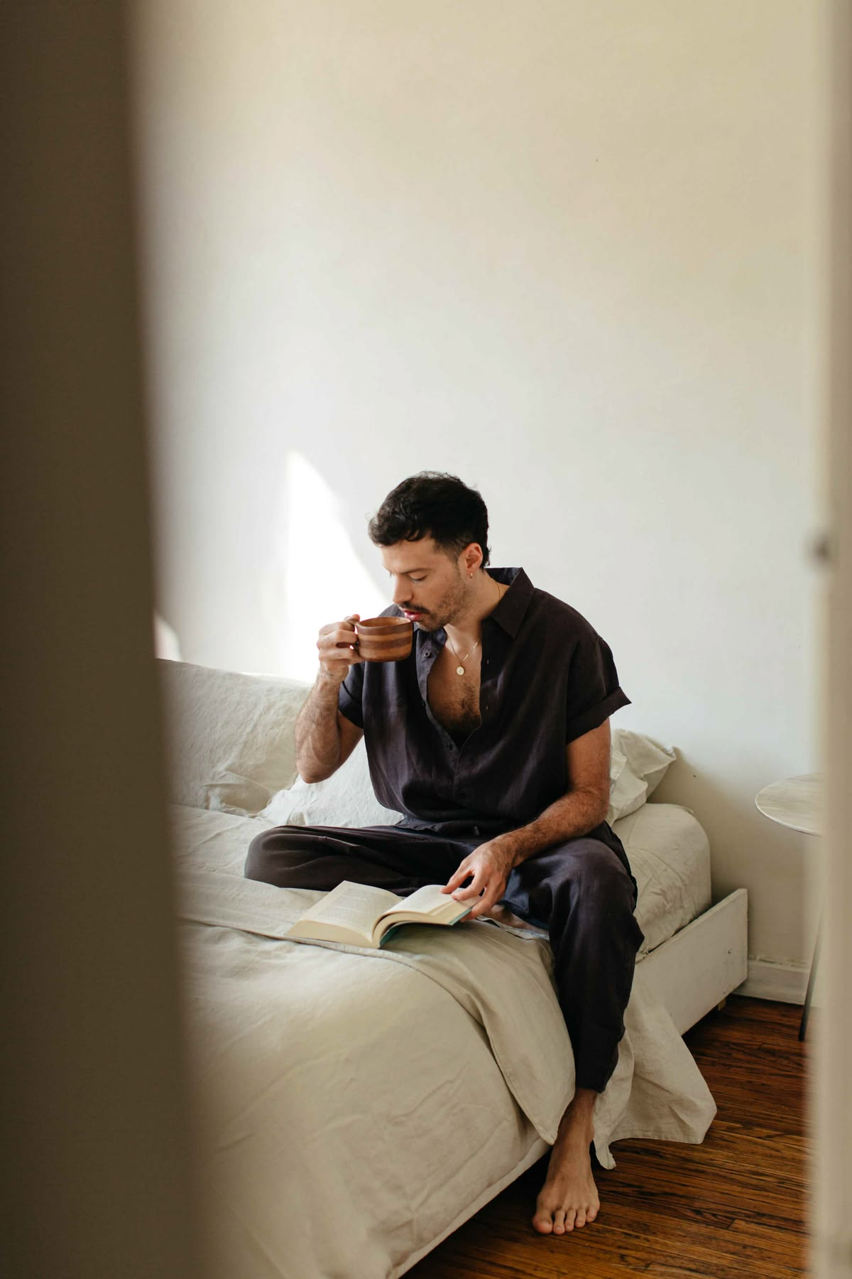 Man sitting cross-legged on bed, drinking from a brown mug and reading a book.