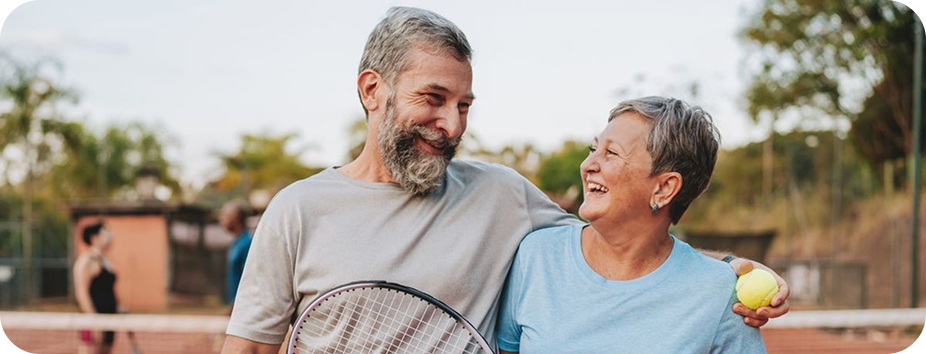Smiling middle-aged couple holding tennis racket and ball on outdoor tennis court with blurred players.