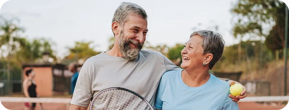 Smiling middle-aged couple holding tennis racket and ball on outdoor tennis court with blurred players.