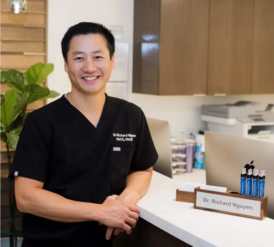Smiling man in black medical scrubs leaning on a reception desk with a nameplate reading Dr. Richard Nguyen.