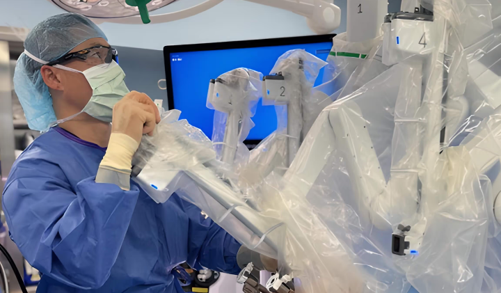 Surgeon in mask and gown operating robotic surgical system covered in plastic drapes.