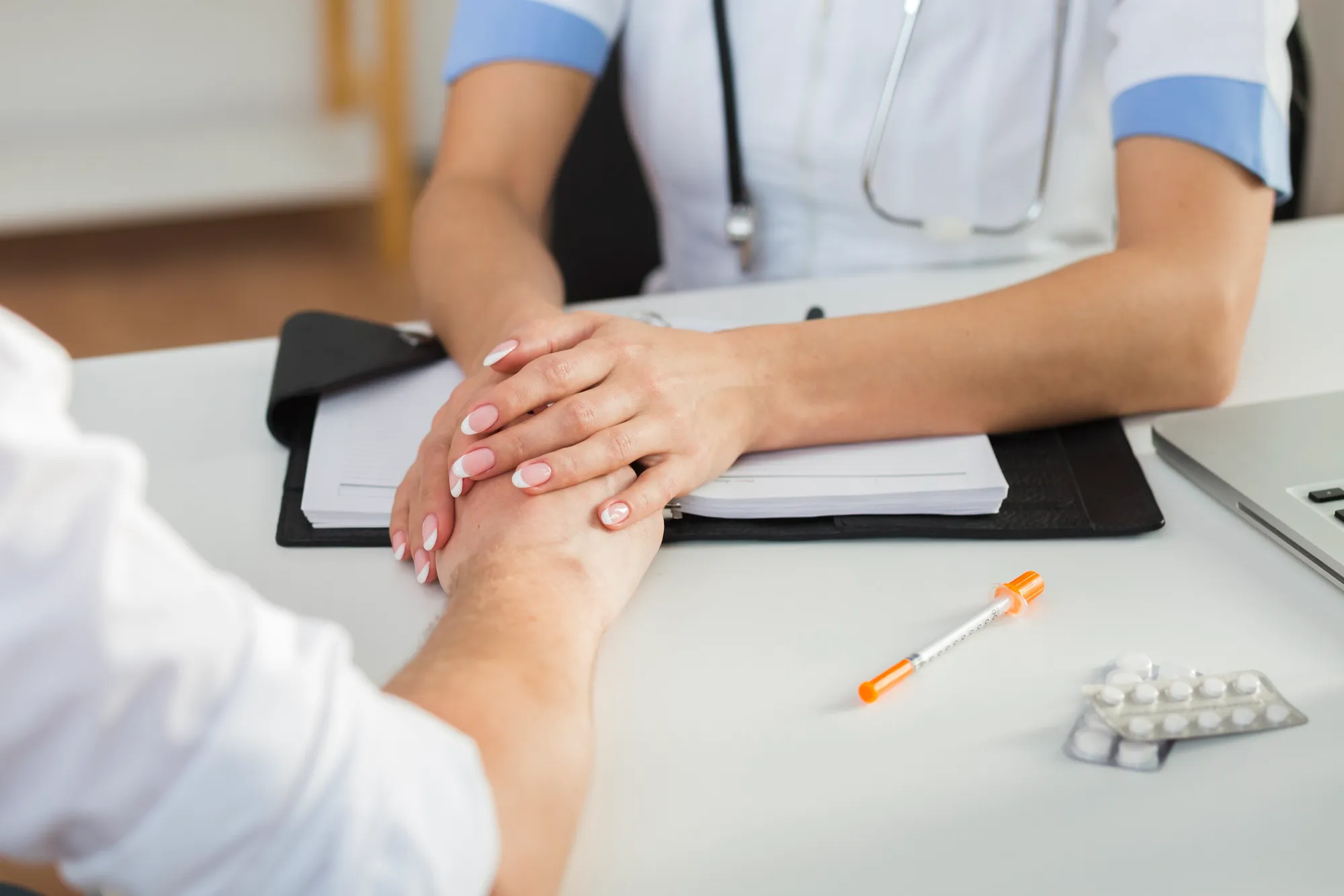 Medical professional holding a patient's hand over a desk with paperwork, pills, and a syringe.