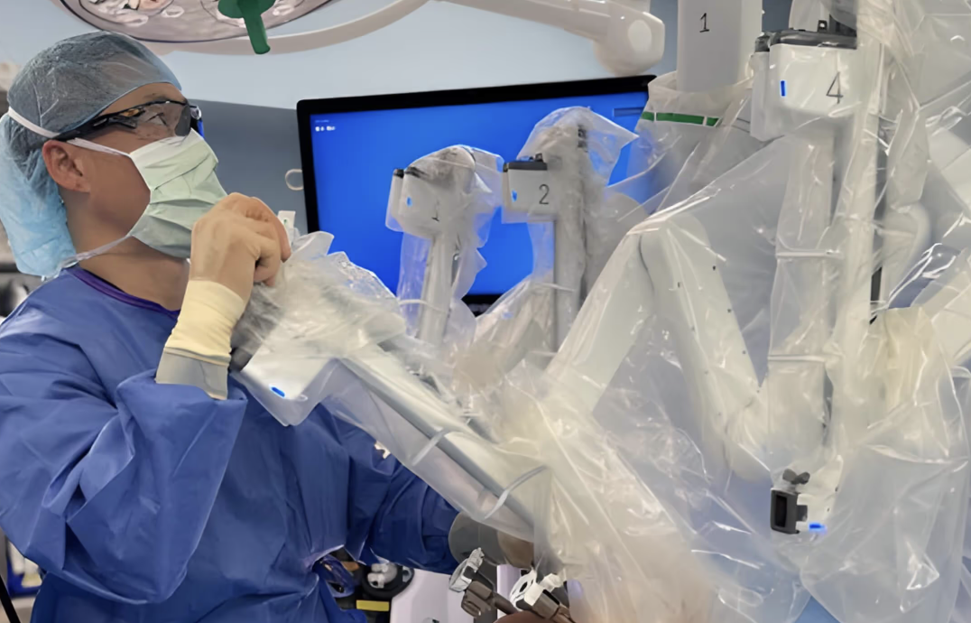 Surgeon in blue scrubs and mask operating a robotic surgical system covered in plastic.