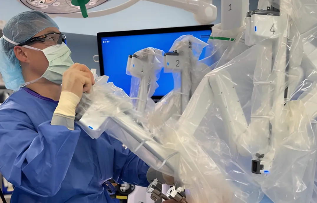 Surgeon in blue scrubs and mask operating a robotic surgical system covered in plastic.