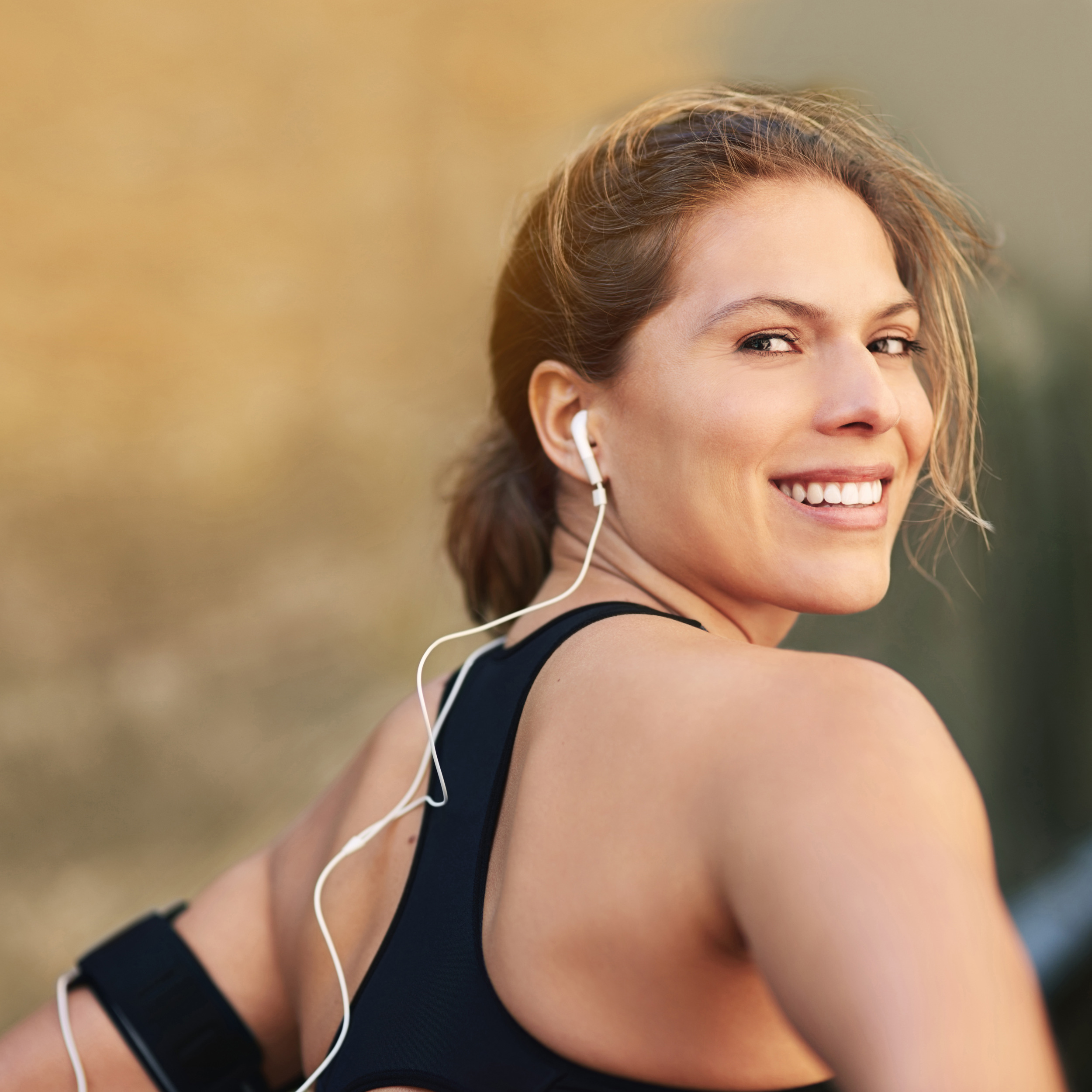 Smiling woman in athletic gear with earphones and armband, outdoors during exercise.