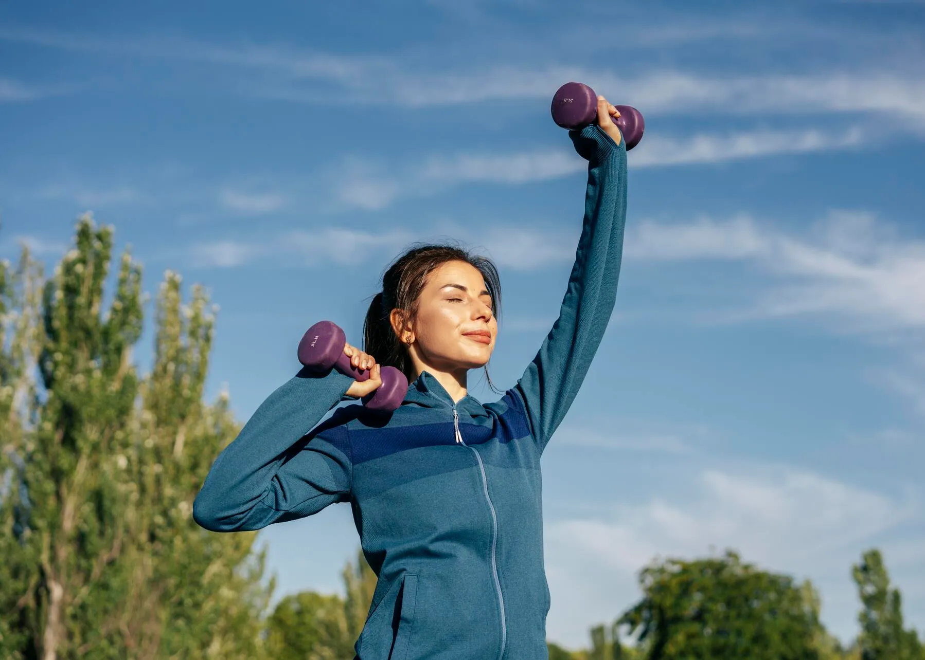 Woman in blue jacket exercising outdoors with purple dumbbells under a blue sky.