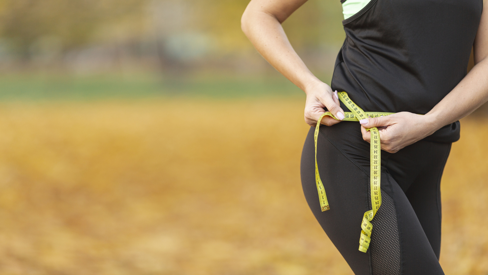 Person measuring their hip with a yellow tape measure outdoors in autumn with fallen leaves.