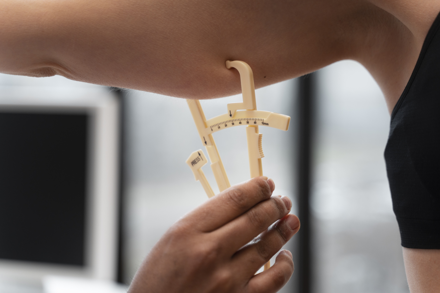 A person measures arm fat thickness with a beige adipometer clamp on the bicep.