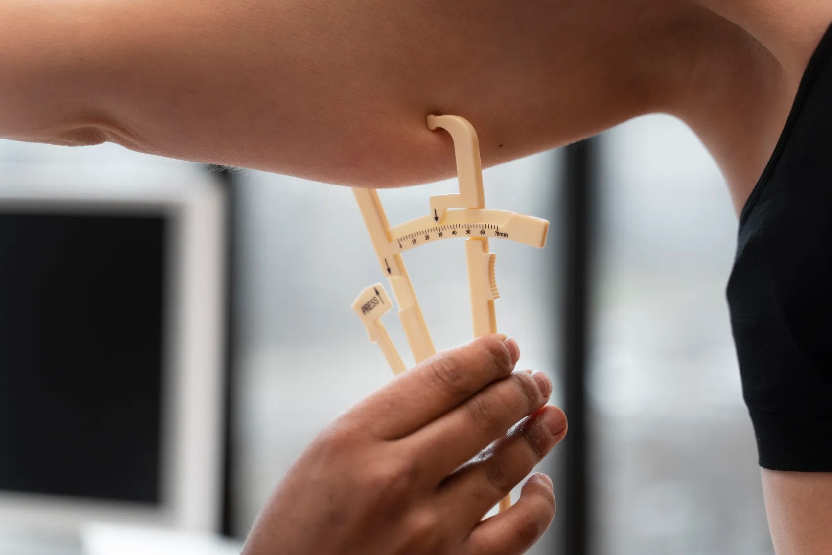 A person measures arm fat thickness with a beige adipometer clamp on the bicep.
