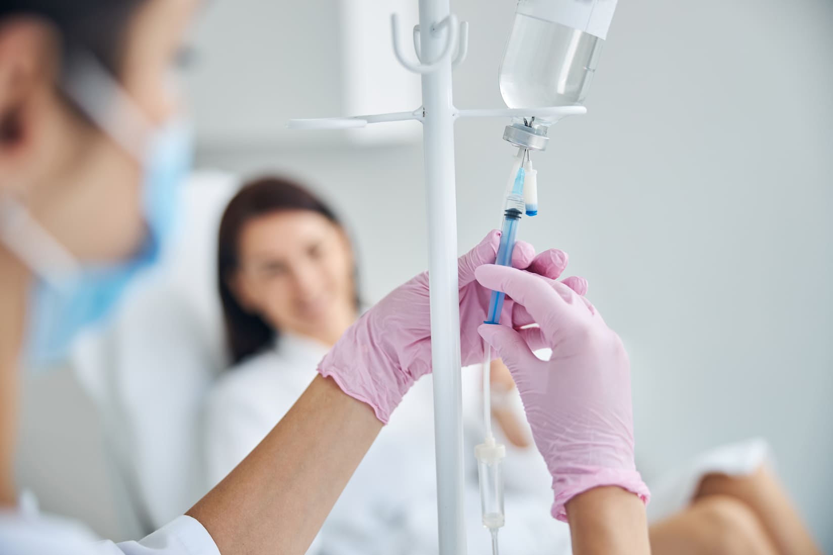 Medical professional in pink gloves adjusting IV drip with patient in the background wearing a mask.