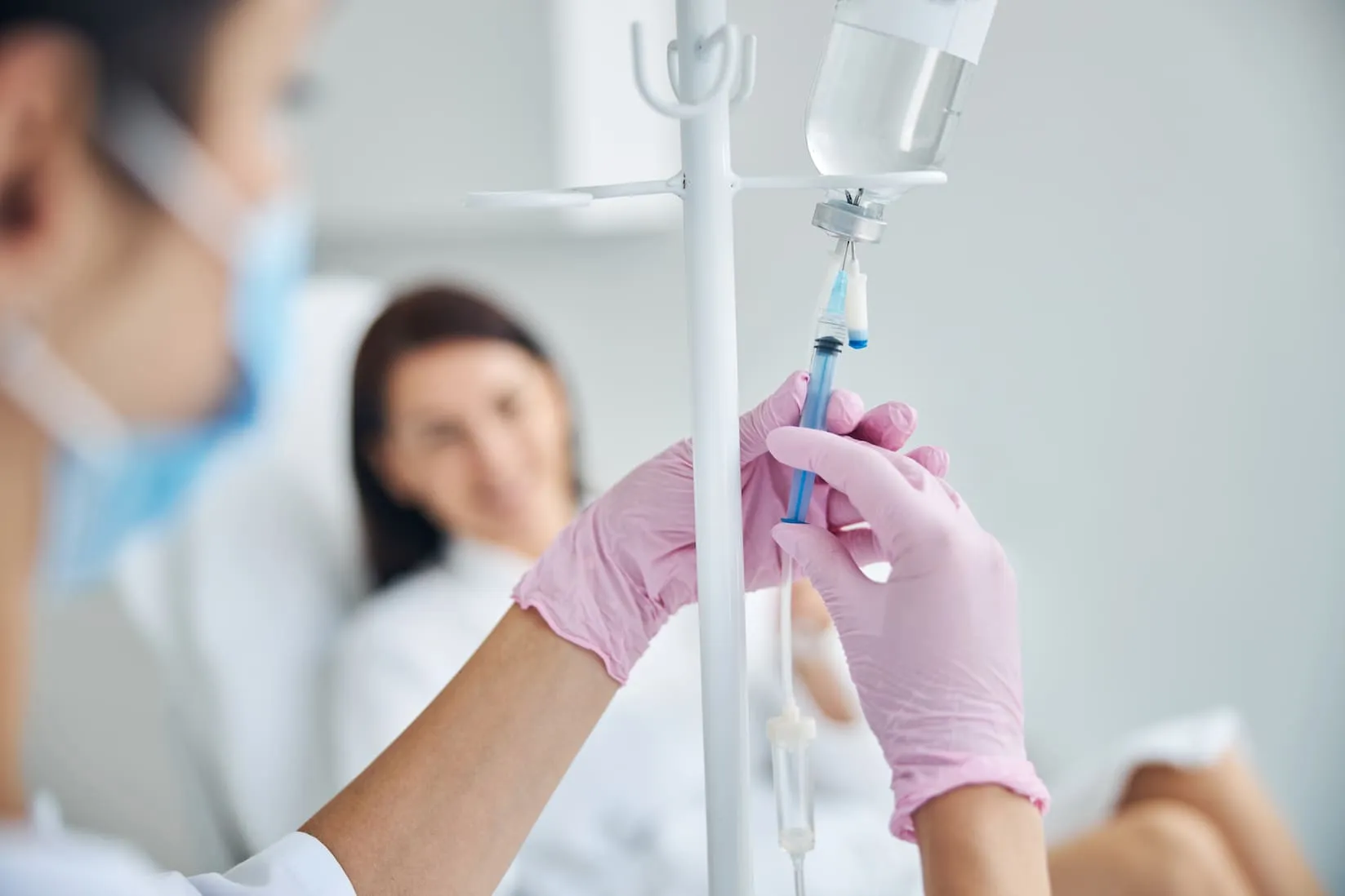 Medical professional in pink gloves adjusting IV drip with patient in the background wearing a mask.