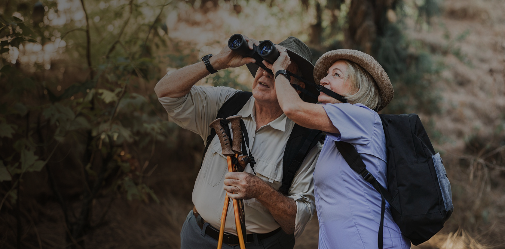 Older couple hiking in the woods, one looking through binoculars, staying active with longevity medicine