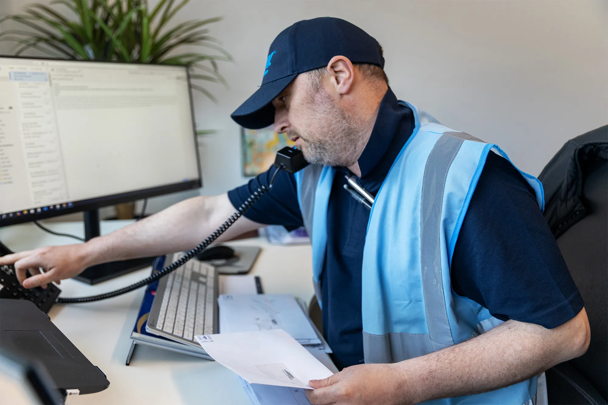 Man in a blue vest and cap talking on a desk phone while holding paperwork and working at a computer.