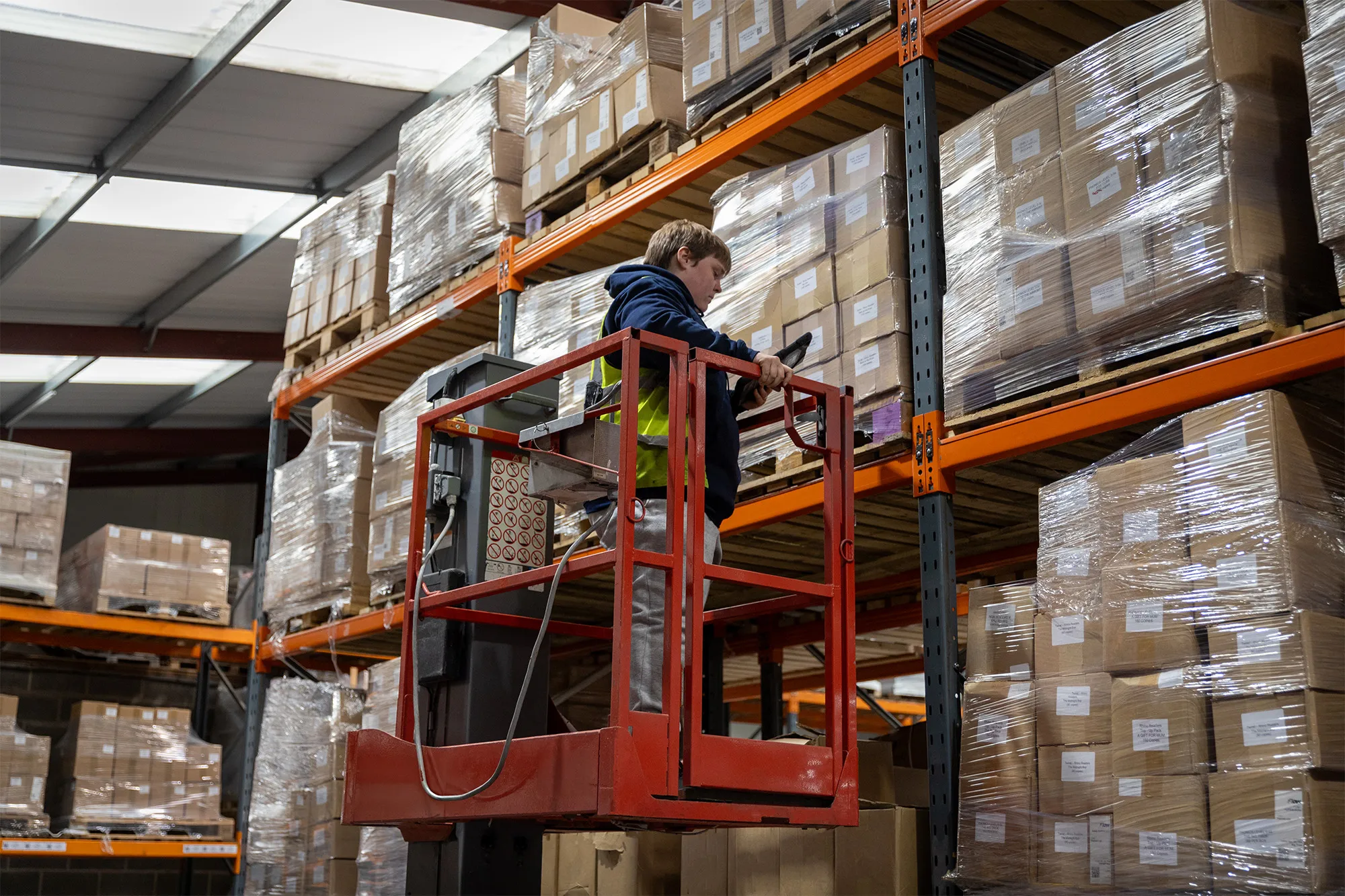Warehouse worker on a red lift scanning barcodes on stacked pallets wrapped in plastic.