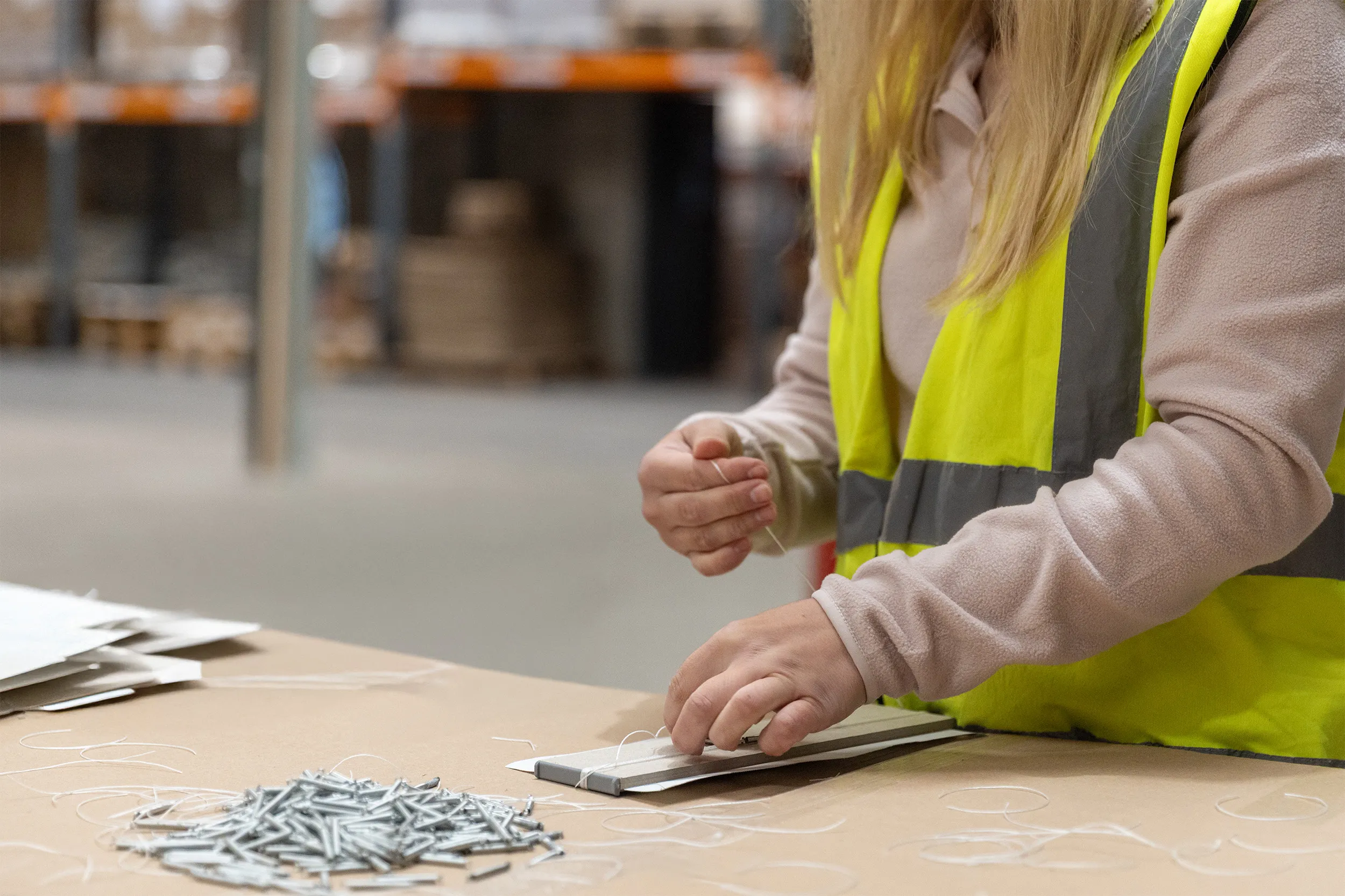 Person in safety vest tying string around stacked flat cardboard pieces on a table with scattered metal rods nearby in a warehouse.