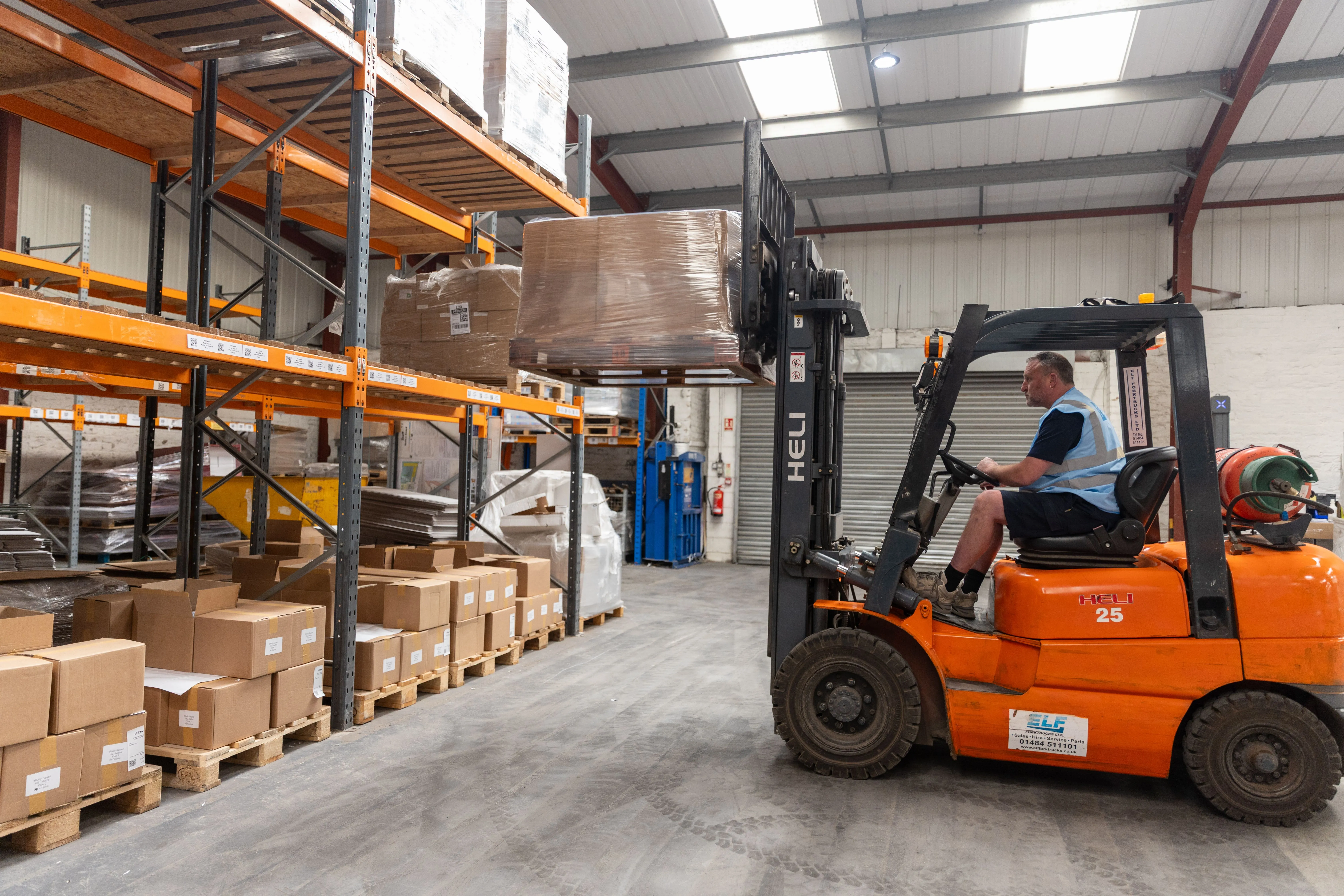Man operating an orange forklift lifting a pallet wrapped in plastic in a warehouse aisle with shelves full of cardboard boxes.