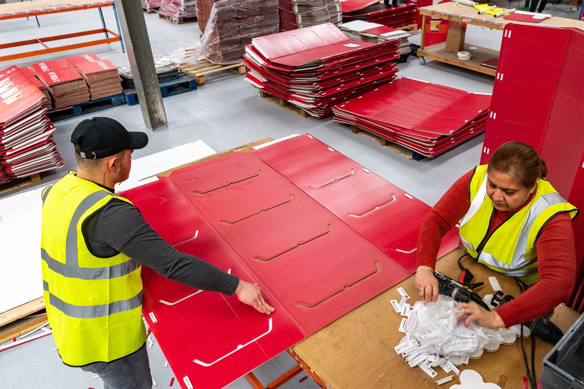 Two workers in high-visibility vests assembling red cardboard sheets in a warehouse filled with stacked cardboard materials.