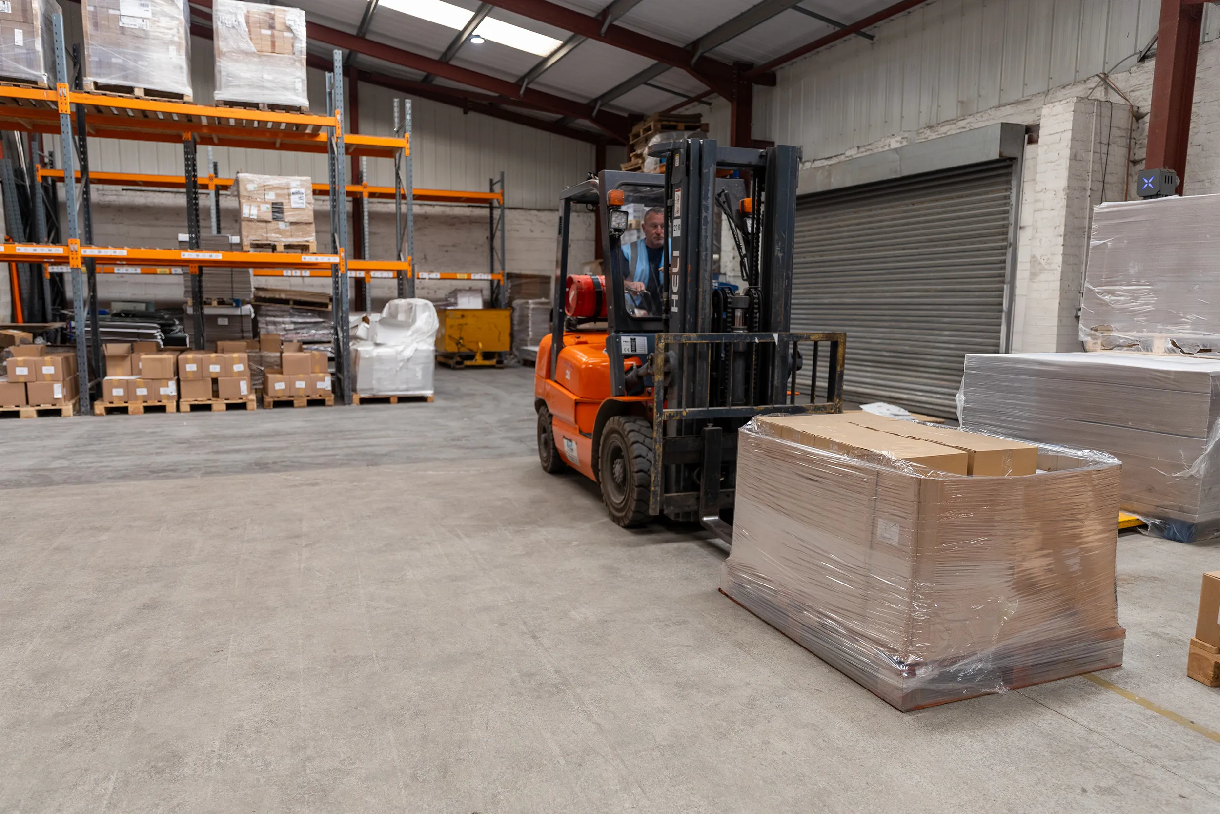 Warehouse worker operating an orange forklift carrying a pallet wrapped in plastic with cardboard boxes.