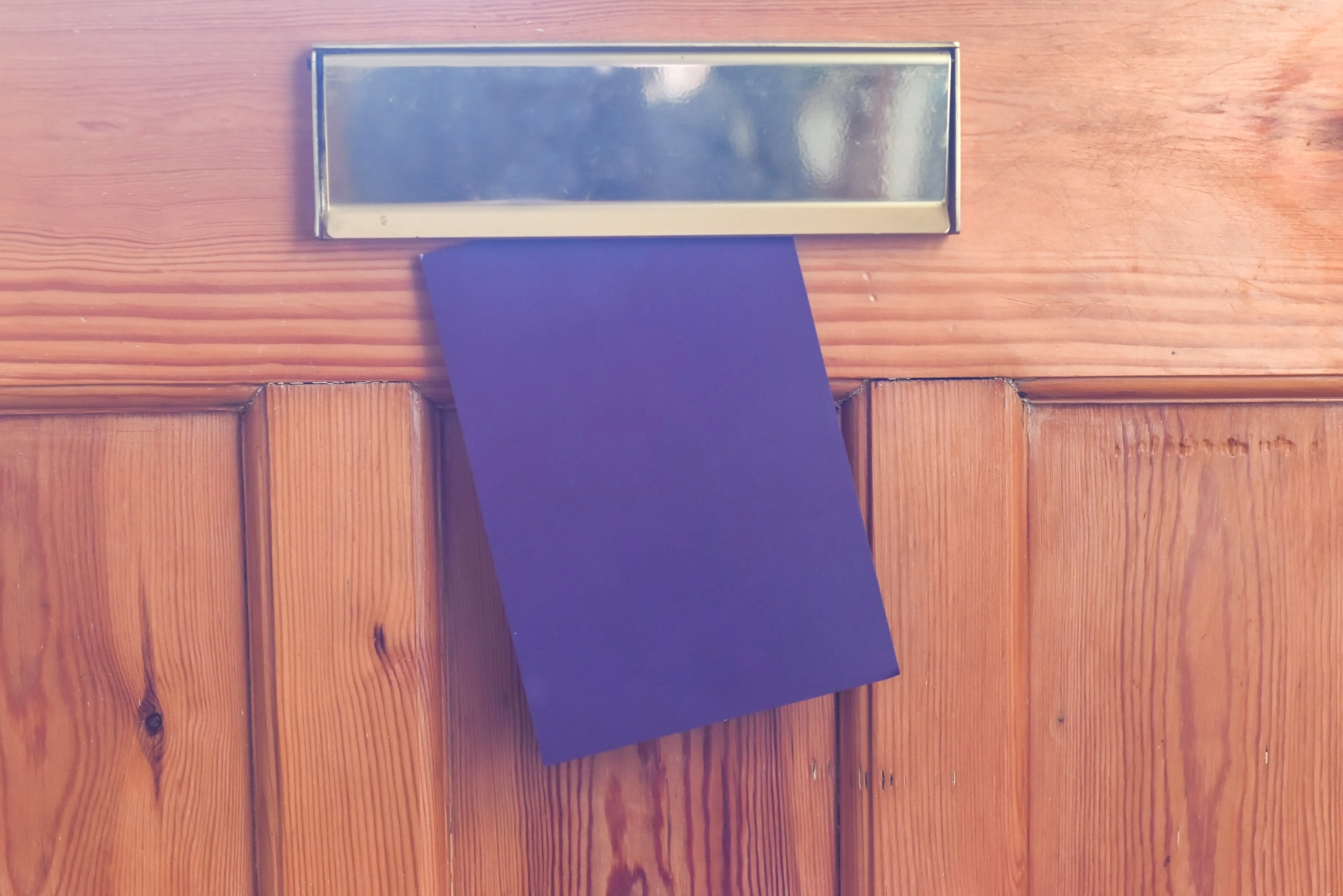 Purple envelope partially inserted into a brass mail slot on a wooden door.