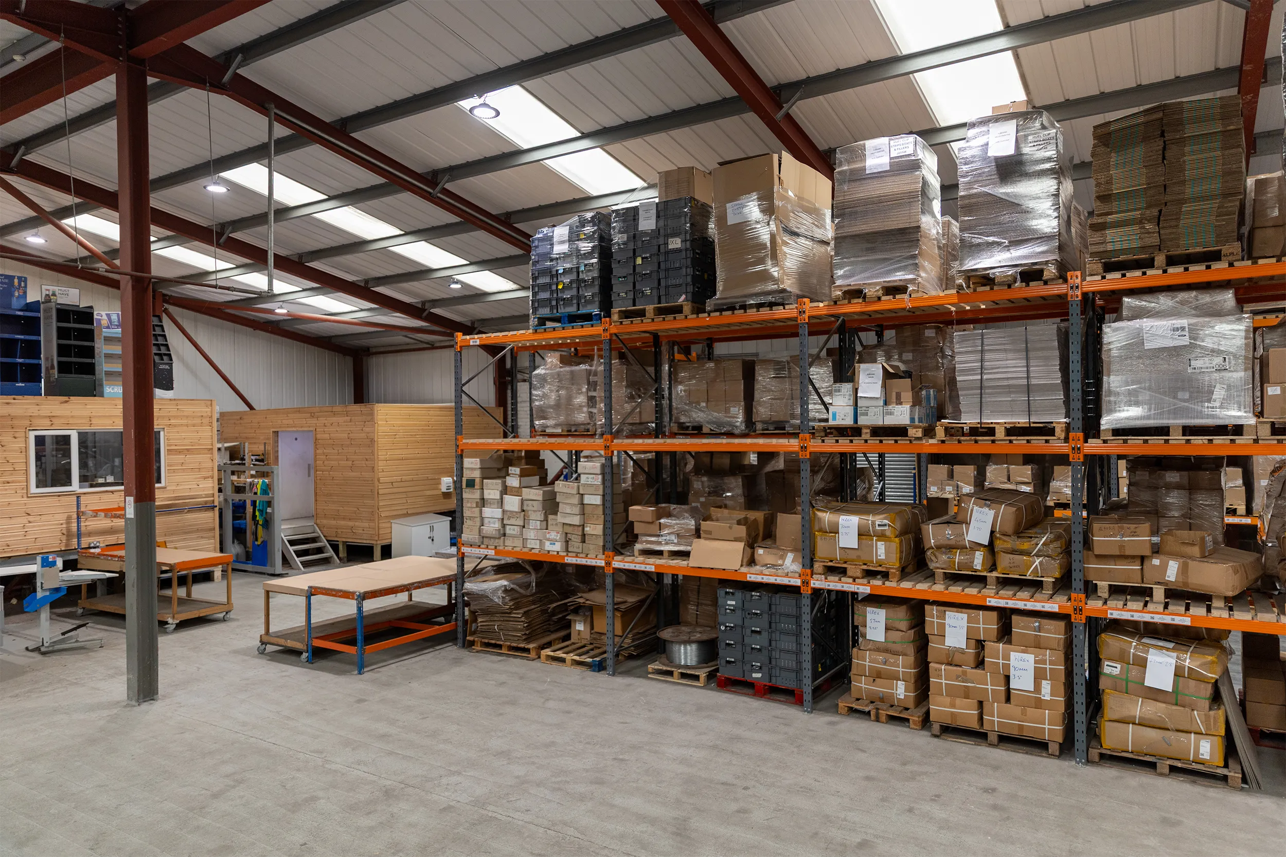 Well-organized warehouse interior with metal shelving holding pallets of boxes and packages, wooden office structures, and worktables.
