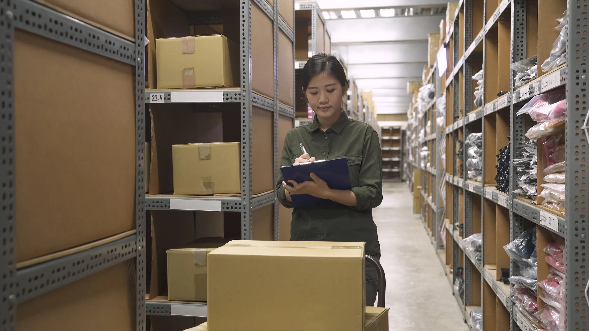 Warehouse worker standing between shelves filled with boxes and plastic packages, writing on a clipboard.