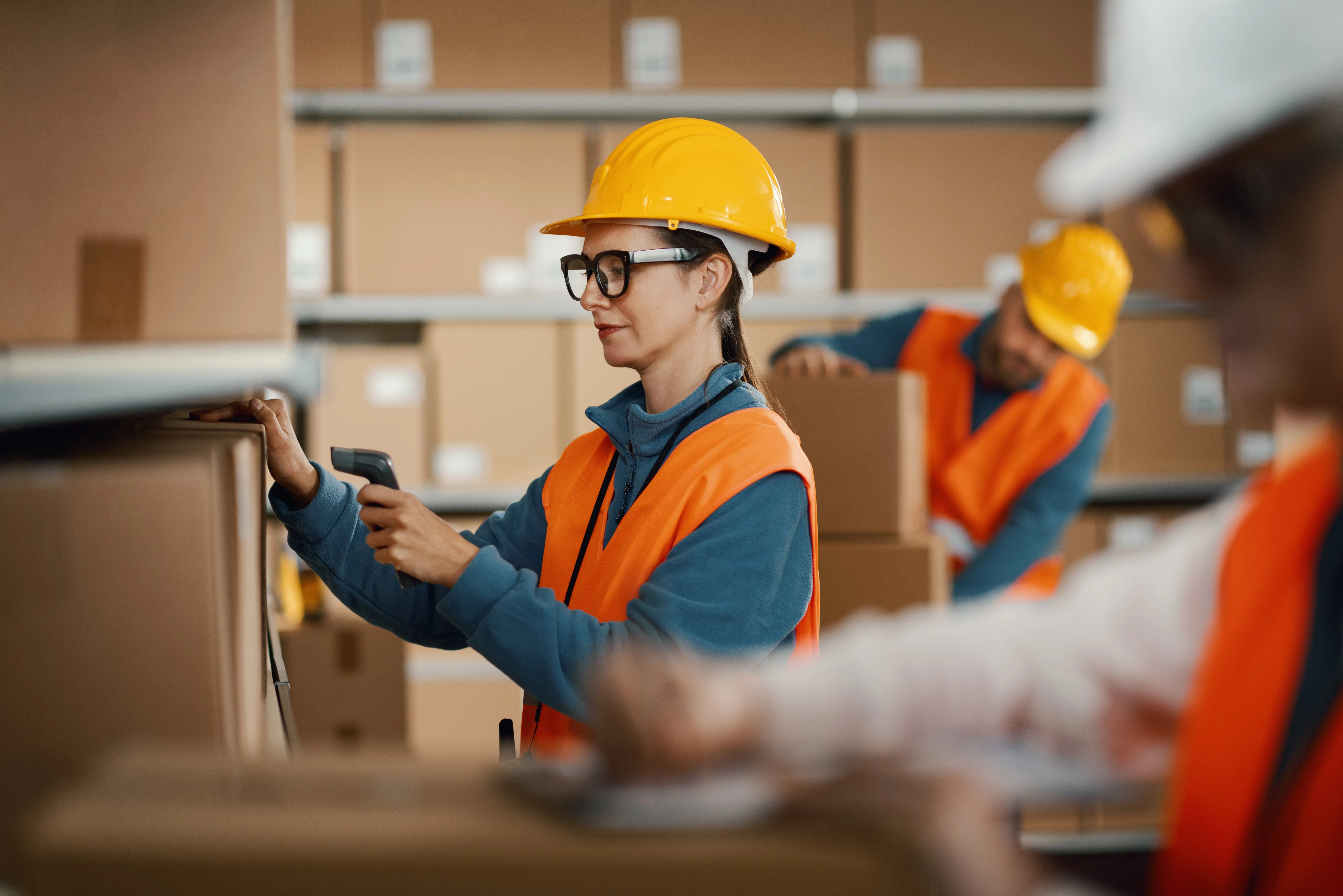 Woman in safety helmet and orange vest scanning a barcode on a box in a warehouse with stacked boxes and coworkers in the background.