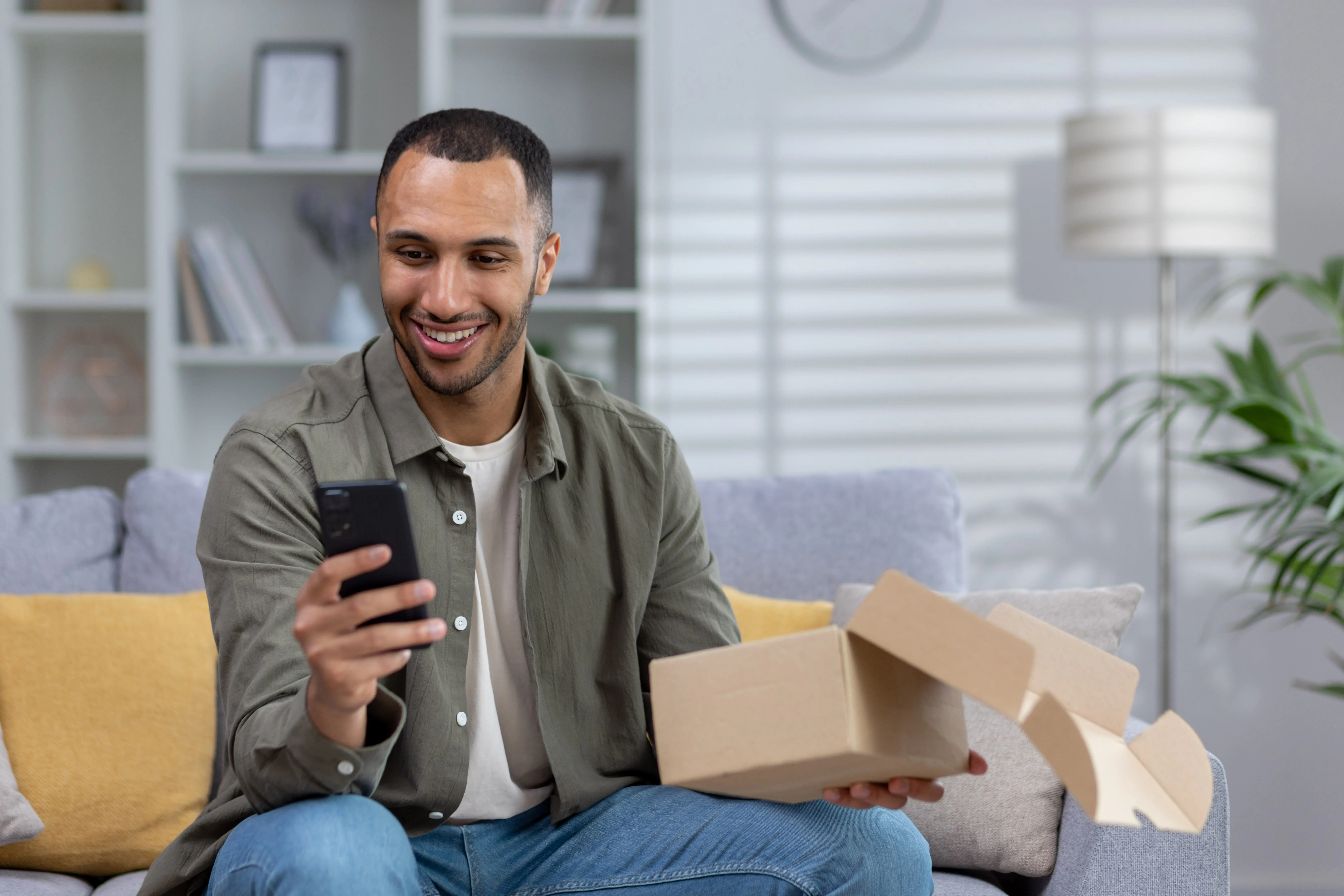 Smiling man sitting on a couch holding an open cardboard box in one hand and looking at his smartphone in the other hand.