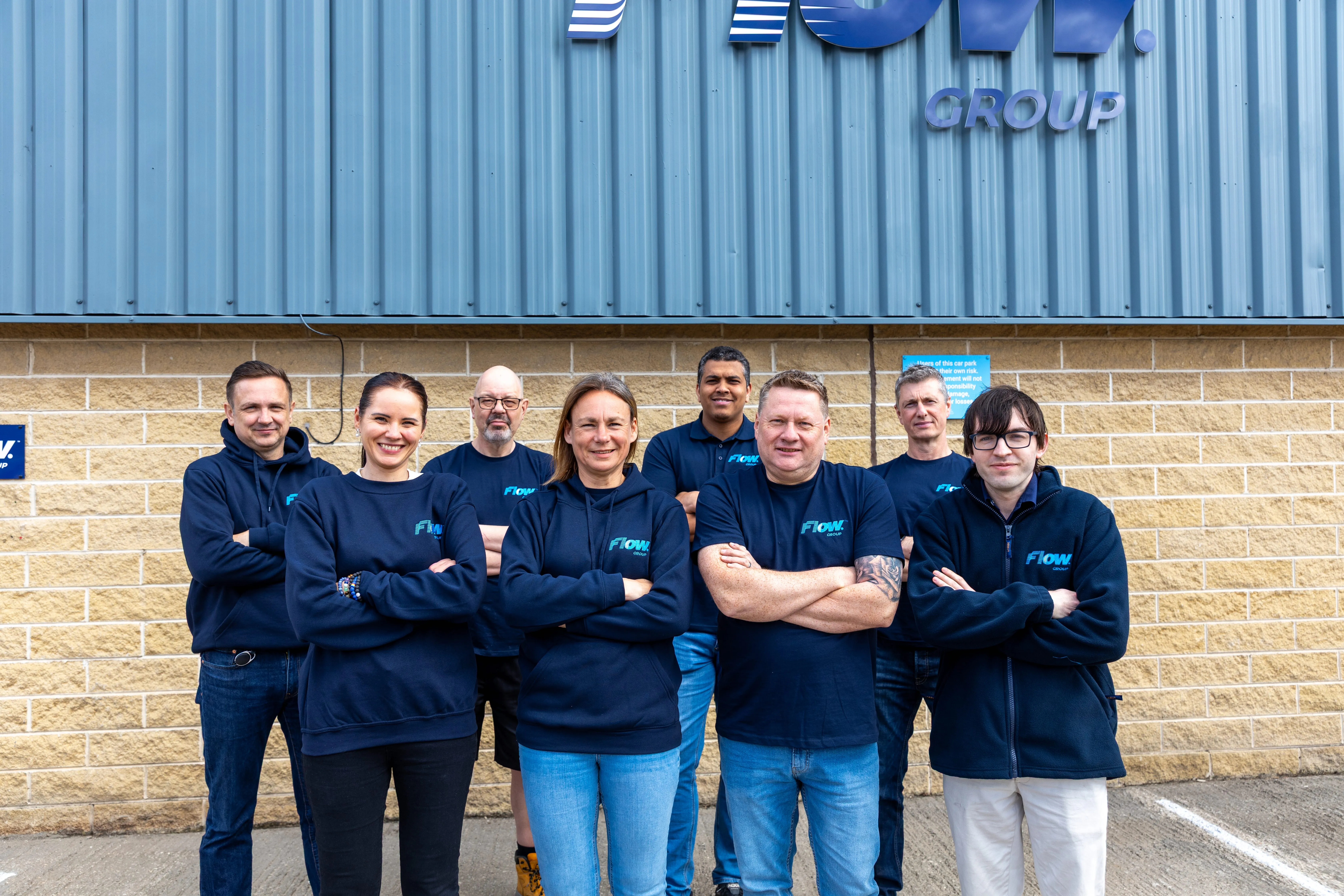 Group of eight people standing outside a building with crossed arms wearing matching navy blue Flow Group shirts and jackets.