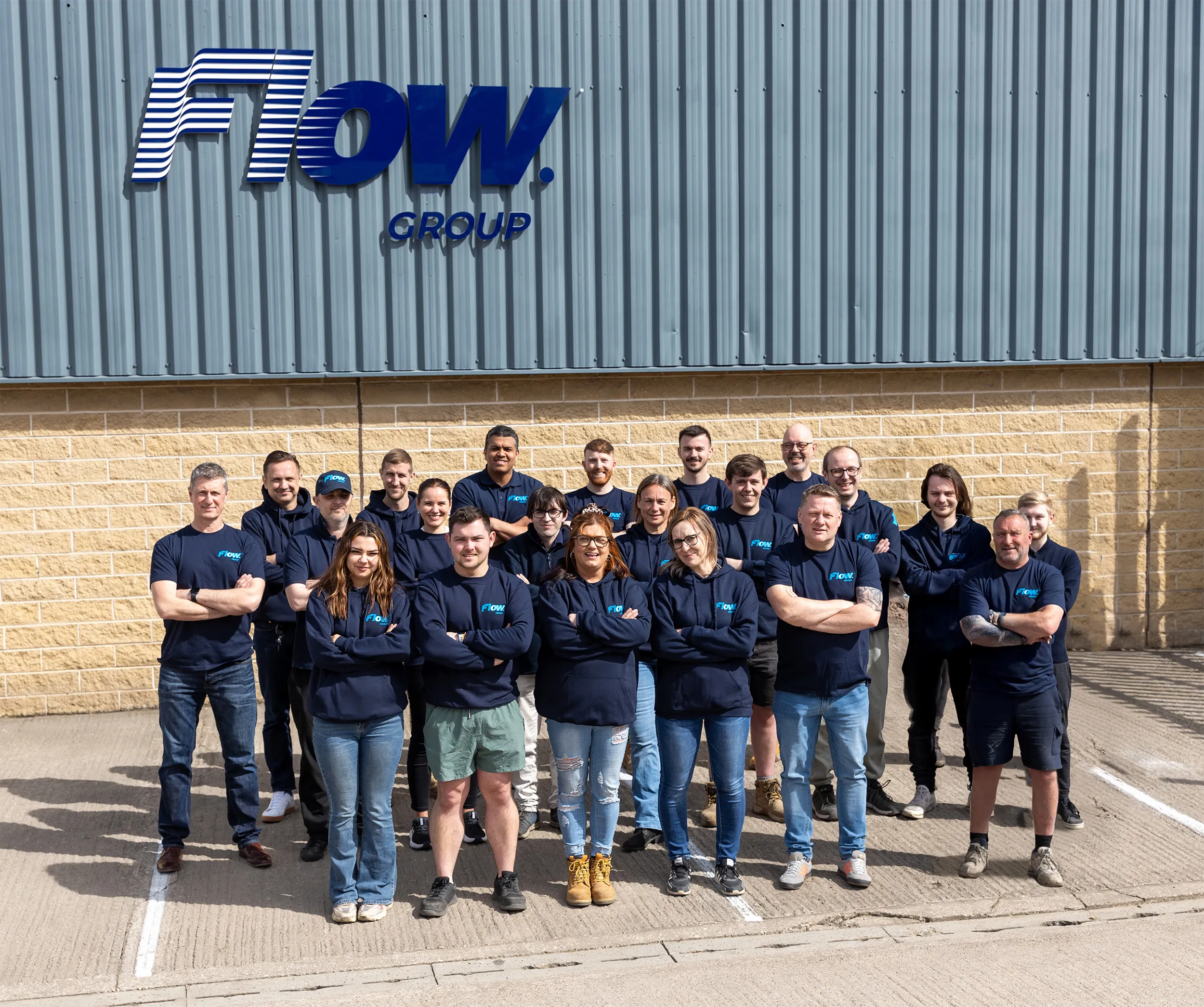 Group of 20 people standing outside a building with a Flow Group sign, wearing matching navy blue shirts or hoodies.